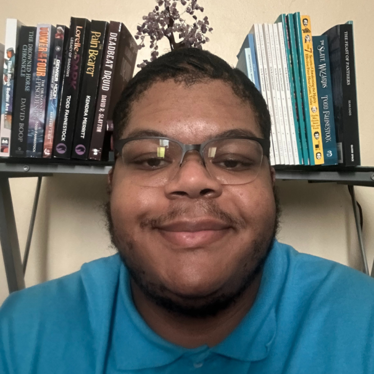 Calvin Shaw headshot - a black man with short black hair smiles softly in front of a bookshelf. He wears rimless glasses and a blue collared shirt.
