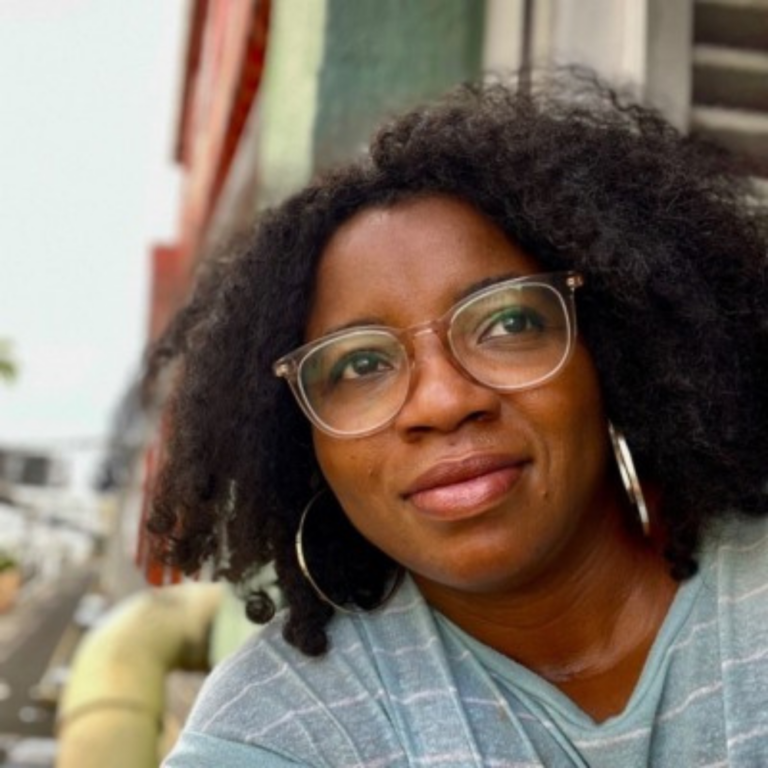 Soni Brown headshot - a black woman with shoulder length hair and wide-rimmed glasses leans out of a window. She is wearing a blue shirt and large silver hoop earrings.