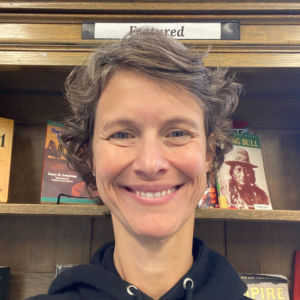 Megan Casimir headshot - a white woman with a wide welcoming smile stands in front of a bookshelf. She has grey-brown hair and wears a black drawstring hoodie.